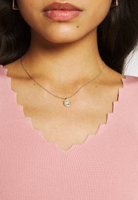 Collana d'argento con un pendente circolare testurizzato indossata sopra una maglia rosa a coste con scollo smerlato.