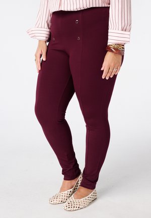 Vrouw die donkerrode aansluitende broek draagt met knoopdetails, een roze gestreepte blouse, gouden en bruine armbanden en crèmekleurige gevlochten platte schoenen.