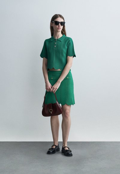 Femme portant un ensemble polo à manches courtes texturé vert et jupe, lunettes de soleil noires, mocassins noirs, tenant un sac à main bordeaux, debout à l'intérieur.