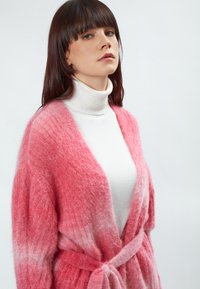 Roze gradient cardigan van een textuur rijke, fluffy stof, met een open voorkant, lange mouwen en een strik taille, over een witte coltrui.