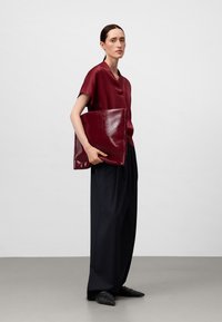 Clutch busta in pelle borgogna tenuta da un modello; top in satin a maniche corte, pantaloni larghi neri e scarpe piatte intrecciate nere.