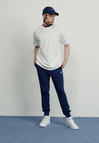 Άσπρο t-shirt Nike, ναυτικά παντελόνια φόρμας και άσπρα αθλητικά παπούτσια. Το μοντέλο φοράει ναυτικό καπέλο. Απλός σχεδιασμός, άνετη εφαρμογή, με λογότυπα Nike στα ρούχα.