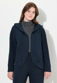 Mörkblå hoodie med mjuk textur, med framficka, fastsydd huva och insydda ärmar, bärs över en svart-och-vitrandig polotröja.