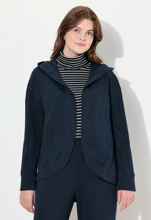 Sudadera azul marino de textura suave, con cremallera frontal, capucha unida y mangas encajadas, usada sobre un cuello de tortuga a rayas en blanco y negro.