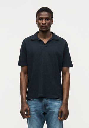 Mlajši moški stoji obrnjeno naprej, oblečen v temno polo majico s kratkimi rokavi in modre kavbojke na enobarvnem svetlem ozadju.