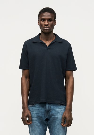 Jeune homme debout de face, portant un polo foncé à manches courtes et un jean bleu sur un fond clair uni.