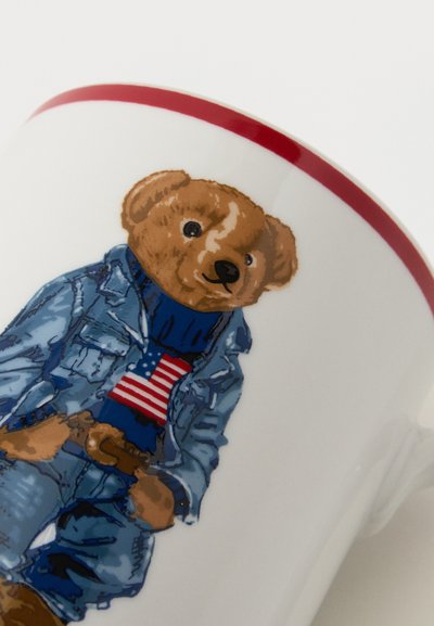 Ursuleț de pluș îmbrăcat într-o jachetă și pantaloni din denim, pulover albastru cu steagul american, imprimat pe o cană albă cu margine roșie.