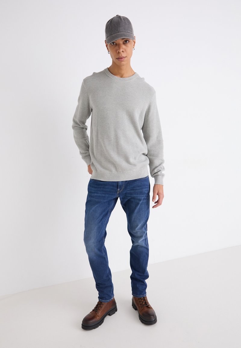 Lichtgrijze gebreide trui, blauwe jeans, bruine veterschoenen en een grijze pet. Model staat met één hand op de heup tegen een effen achtergrond.