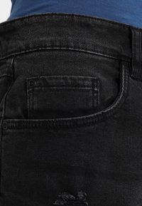 Jeans de mezclilla negra con un parche desgastado, que presentan un bolsillo trasero y detalles de costura estándar. Cintura visible y textura de la tela.