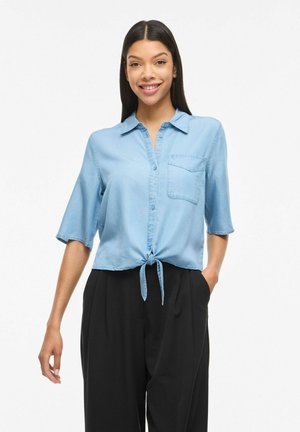 Kobieta z długimi ciemnymi włosami, ubrana w jasnoniebieską koszulę wiązaną z przodu i czarne szerokie spodnie, uśmiechająca się z jedną ręką w kieszeni.