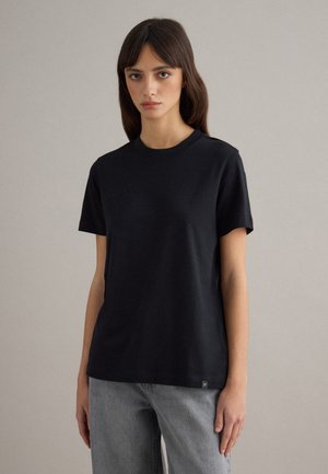 Junge Frau trägt ein schlichtes schwarzes T-Shirt mit kurzen Ärmeln und hellgraue Jeans, steht vor einem neutralen Hintergrund.