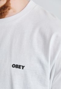 Fehér pamut póló, kerek nyakkivágással. Bal melle részén fekete "OBEY" logóval. Simább textúra, egyszerű dizájn.