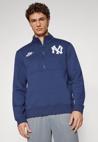 Námornícky modrý pulóver so zvýšeným golierom, zipsovým zapínaním, bielym logom Nike a bielym emblémom New York Yankees na hrudi.