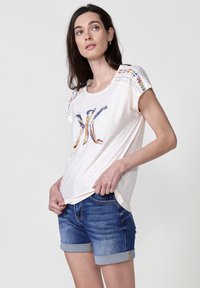 T-shirt blanc à manches courtes avec un design brodé multicolore et des accents à clous sur les épaules, associé à un short en denim.