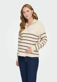 Suéter beige con rayas horizontales marrones, con cuello con cremallera y textura acanalada. Combinado con jeans azul oscuro.
