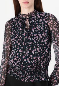 Blusa nera a fiori con maniche lunghe trasparenti, vita arricciata e dettagli con fiocco al collo. I motivi includono fiori rosa e blu su uno sfondo scuro.