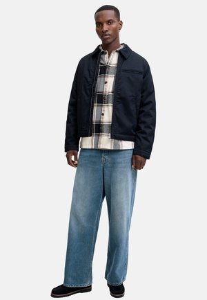 Uomo in piedi che indossa una giacca blu scuro sopra una camicia a quadri beige e nera, jeans blu a gamba larga e scarpe nere, su uno sfondo semplice.