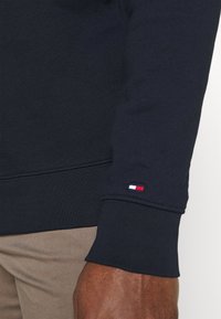 Felpa a maniche lunghe blu navy con polsino e piccolo logo a bandiera rosso, bianco e blu sul polso, indossata sopra dei pantaloni beige su un braccio di carnagione scura.
