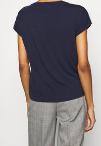 Osoba s médii pleti, oblečená v námořnickém tričku s krátkým rukávem a šedých kostkovaných kalhotách, zobrazená zezadu, stojící na bílém pozadí.