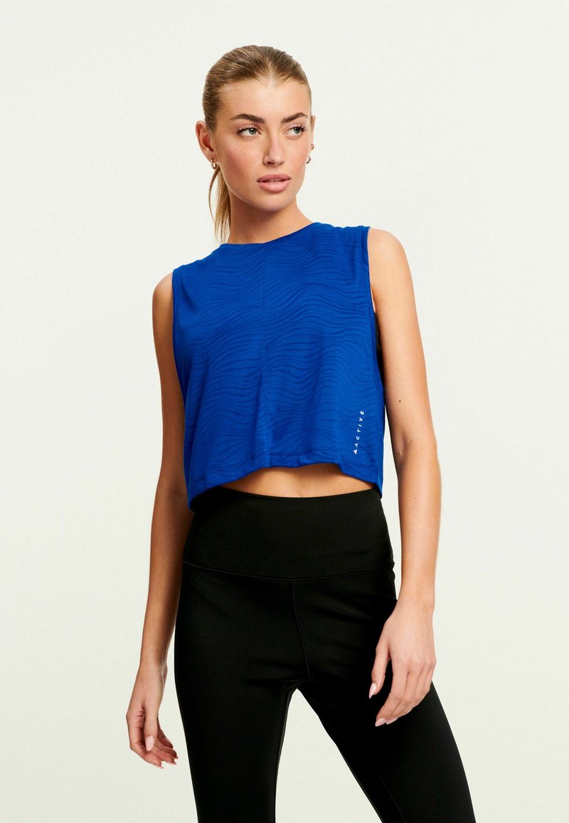 Blå ærmeløs cropped top med tekstureret bølgemønster og subtilt mærkedetalje, parret med højtaljede sorte leggings.