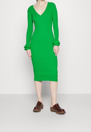 Frau trägt ein leuchtend grünes, langärmeliges knielanges Kleid mit gerafften Bündchen und braune Slip-On-Clogs mit Holzsohle.