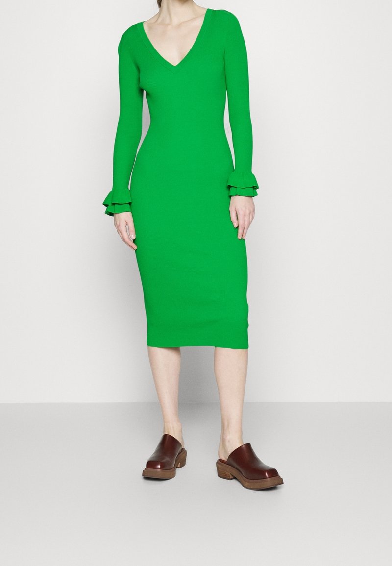 Femme portant une robe verte vif à manches longues, longueur genoux, avec des poignets à volants et des sabots marron à enfiler avec une semelle en bois.