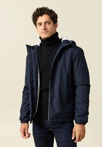 Giacca impermeabile blu navy con cappuccio, dotata di polsini elasticizzati e foderata in morbido pile. Indossata sopra un maglione nero a trecce.