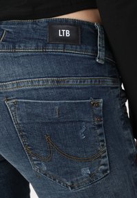 Tmavě modré džíny z denimu s koženou nášivkou označenou "LTB," s žlutým prošíváním, kapsami a poškozenými detaily poblíž kapes.