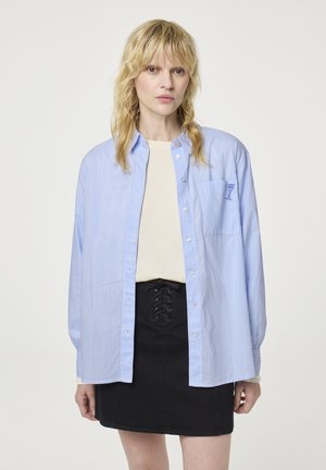 Lichtblauw gestreept overhemd met lange mouwen, voorzien van een borstzak en een klein grafisch ontwerp. Gecombineerd met een zwarte mini-jurk.