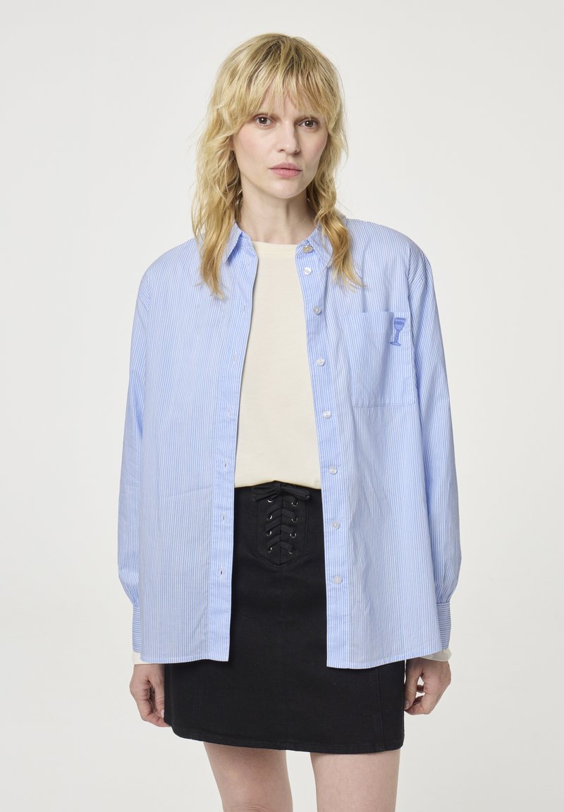 Light blue striped button-up shirt with long sleeves, featuring a chest pocket and a small graphic design. Paired with a black mini skirt.