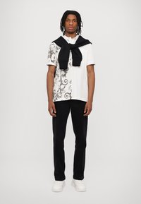 Balts T-krekls ar melnu paisley rakstu vienā pusē, savienots ar melnām biksēm un baltām kedām. Ap pleciem ir uzmests melns džemperis.