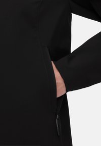 Zwarte softshell jack met een ritszak aan de zijkant. Heeft een gladde textuur en een gestroomlijnd design. Minimale hardware zonder zichtbare accenten.
