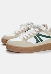 Sneakers z kremową cholewą z zamszu, białymi skórzanymi panelami, zielonymi akcentami i gumową podeszwą. Posiadają sznurówki i wzmocnione szwy.
