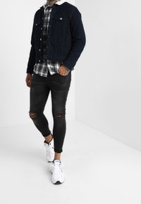 Marinblå denimjacka med vit krage, svart-vit rutiga skjorta under, slitna svarta smala jeans och vita träningsskor.