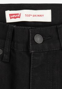 Fekete farmer Levi's 510 skinny farmernadrág, piros szöveget tartalmazó címkével. Kiemelkedő részletek közé tartoznak a fekete fém gombok és a varrási részletek.