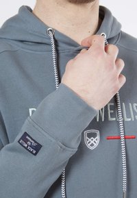 Haut à capuche gris avec un cordon de serrage, doté d'un cordon tressé noir et blanc contrastant, d'un texte brodé et d'un petit patch logo sur la manche.