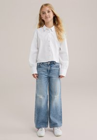 Niña joven con cabello largo y rubio, vistiendo una camisa blanca de botones, jeans de pierna ancha y rasgados, y zapatillas blancas, de pie contra un fondo liso.