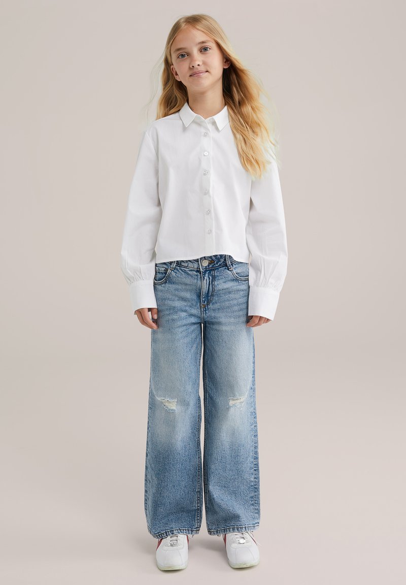 Niña joven con cabello largo y rubio, vistiendo una camisa blanca de botones, jeans de pierna ancha y rasgados, y zapatillas blancas, de pie contra un fondo liso.