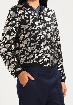 Musta vetoketjullinen bomber-takki, jossa hopeinen kukkakirjonta, käytetty sinisen kauluspaitaan ja tummansinisten housujen päällä.