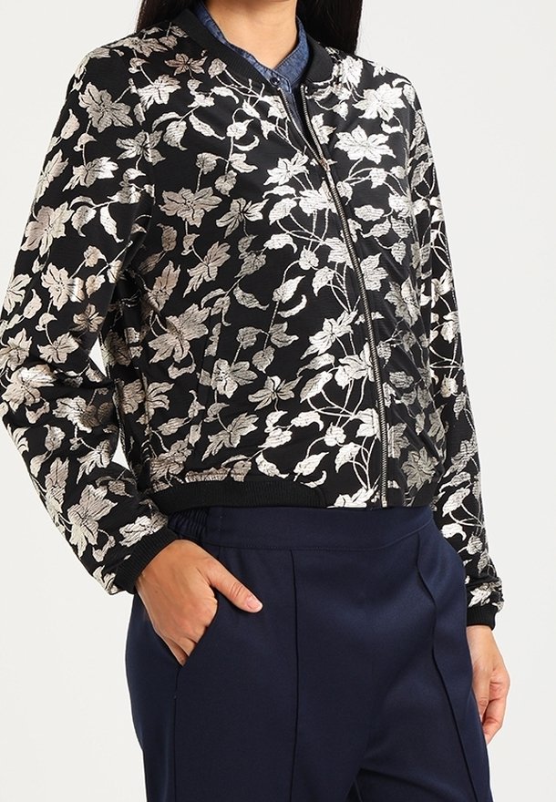 Veste bomber noire zippée avec broderie florale argentée portée sur une chemise bleue à col et un pantalon bleu marine.