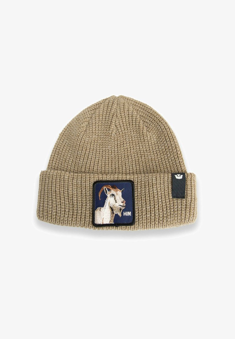 Gorro de punto beige con puño doblado, que presenta un parche de cabeza de cabra blanca sobre un fondo azul marino y una pequeña etiqueta negra en el lateral.