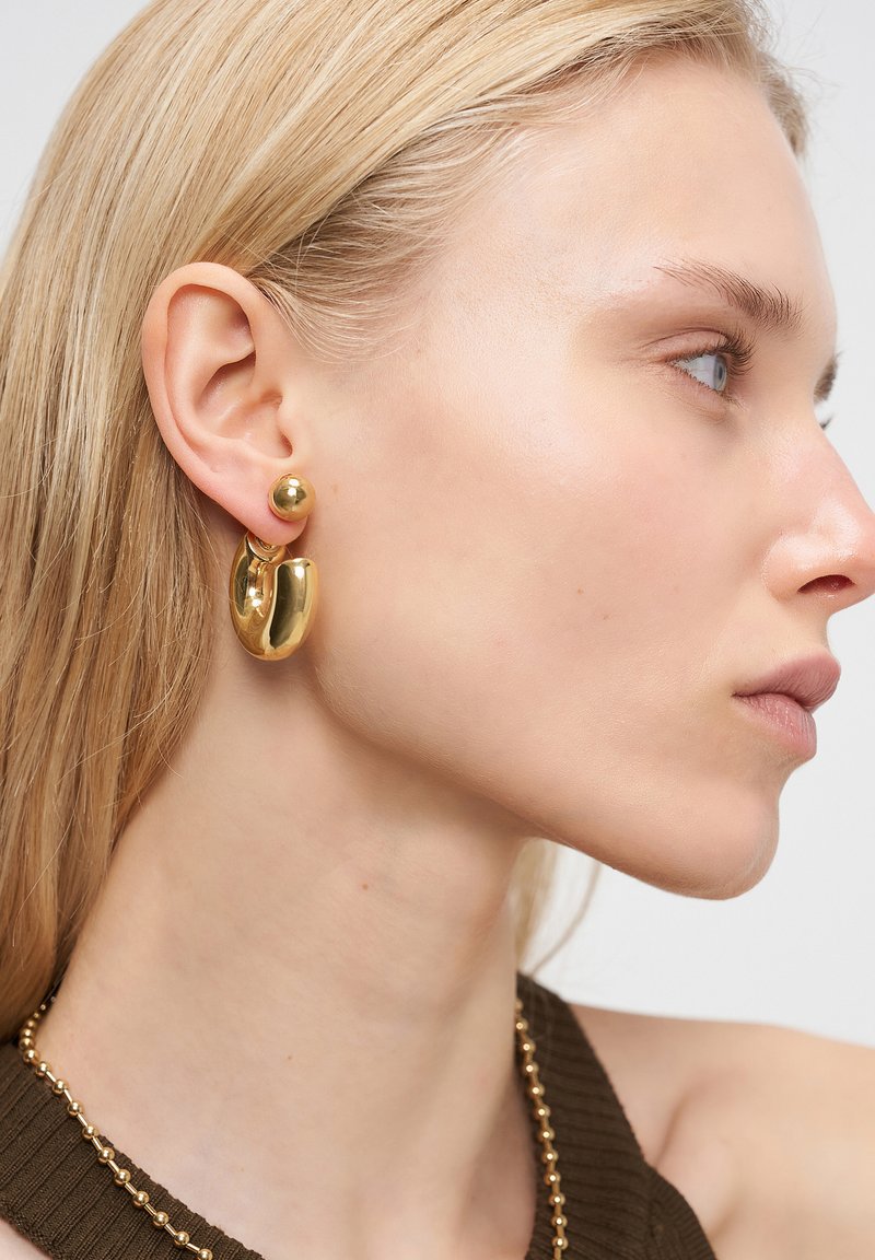 Goldfarbene metallische Ohrringe mit einem runden Design, das eine kleine Kugel und eine größere unregelmäßige Ringsch_form umfasst. Glatte, reflektierende Oberfläche.