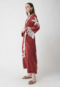 Robe en coton rouge de style kimono avec des franges blanches et une broderie de feuilles, dotée de larges manches et d'un ourlet effiloché. Associée à des sandales plates.