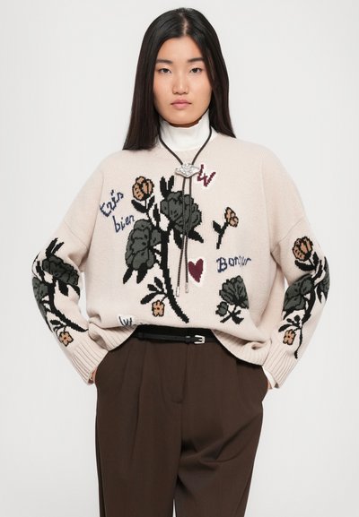 Lichtroze gebreide trui met bloemenborduursel in donkergroen, oranje en blauw, met een brede halslijn en ribgebreide manchetten, gedragen over bruine broek.