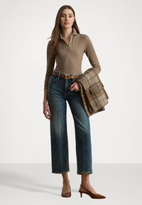 Top de mangas longas com riscas, castanho, e colarinho, jeans de cintura alta azuis, e mules castanhos de salto. Um blaser bege com padrão quadriculado está pousado sobre um braço.