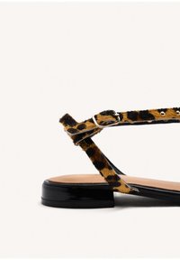 Sandaña plana con estampado de leopardo y una tira de charol negro, que presenta una hebilla metálica ajustable y una plantilla suave en color beige. Tacón bajo.