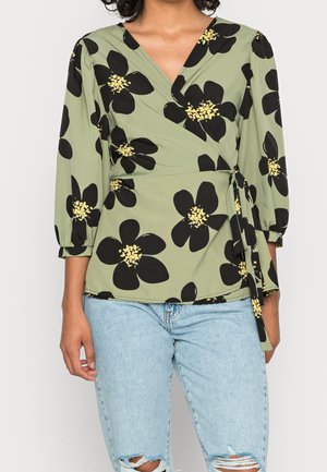 Grøn blomsterwrap-top med sorte blomster og gule centre. Lange ærmer med rynkedetaljer. Kombineret med slidte lyseblå jeans.
