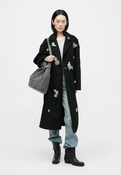 Črn plašč s cvetličnimi vezeninami, dolga silhueta, ovratnik z rezi, v kombinaciji s sivo torbo, svetlim puloverjem in ohlapnimi džinsi. Črne gležnjarje.