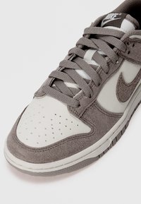 Pilkai suknelės sportbačiai su balta odine pirštų sienele, perforuotu dizainu, pilkomis raištelėmis ir tekstūruotu guminio padu. Nike logotipas ant liežuvėlio.