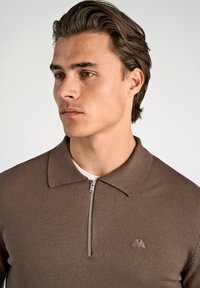 Brun zip-neck tröja gjord av ett texturerat tyg, med en krage och en liten broderad logotyp på bröstet.